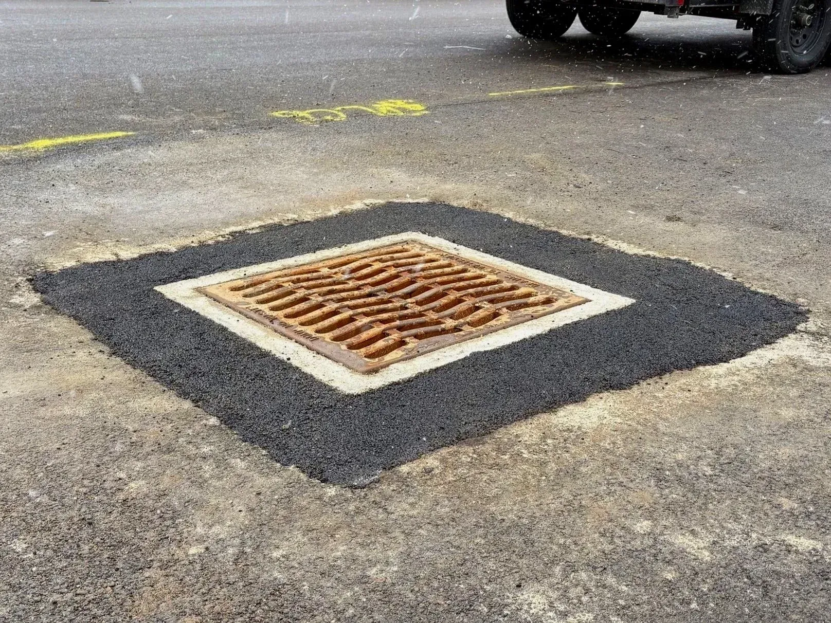 A square storm drain grate in asphalt. A rectangular patch of black asphalt surrounds the drain.