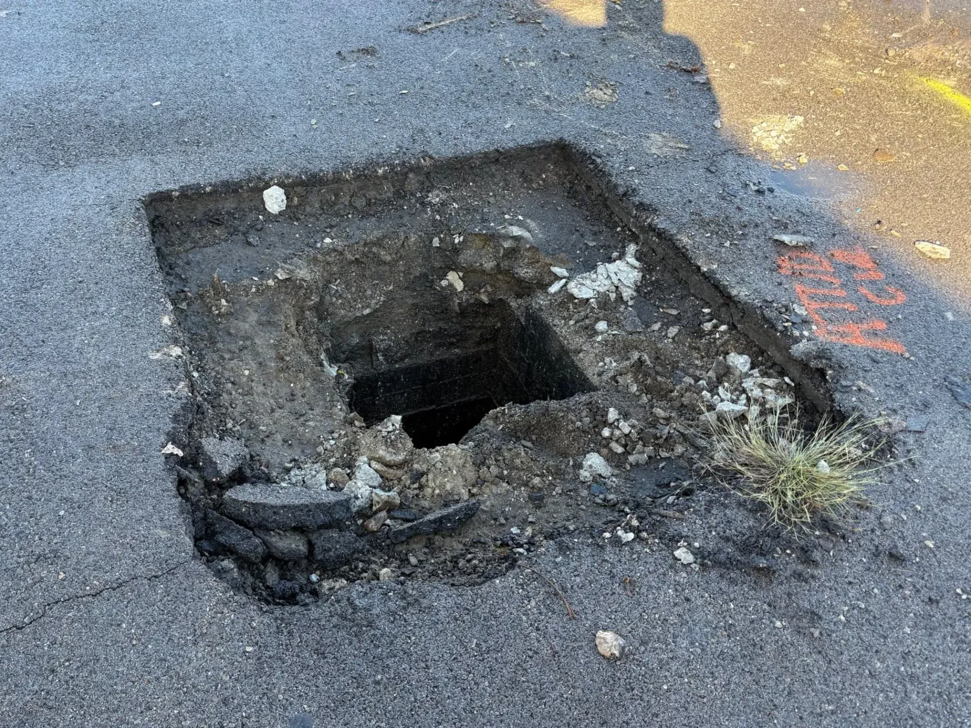 A square hole in asphalt pavement with a dark void, exposing the ground beneath.