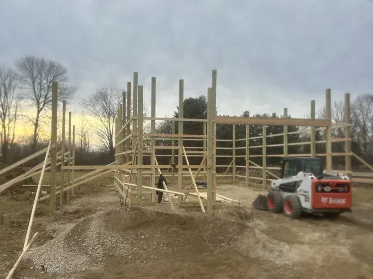 Construction site: wooden barn frame partially built, small Bobcat, overcast sky.