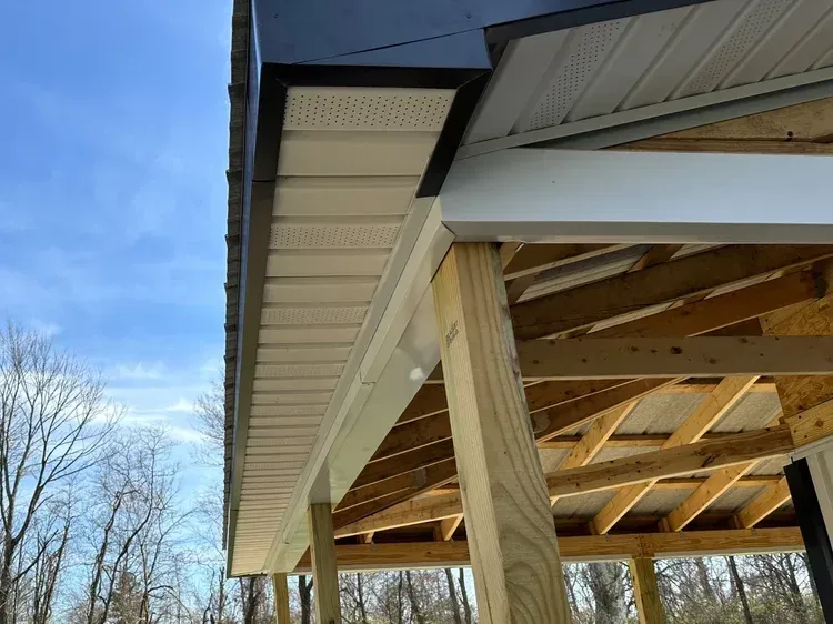 Construction of an outdoor shelter with a white soffit, supported by wooden beams. Blue sky visible.
