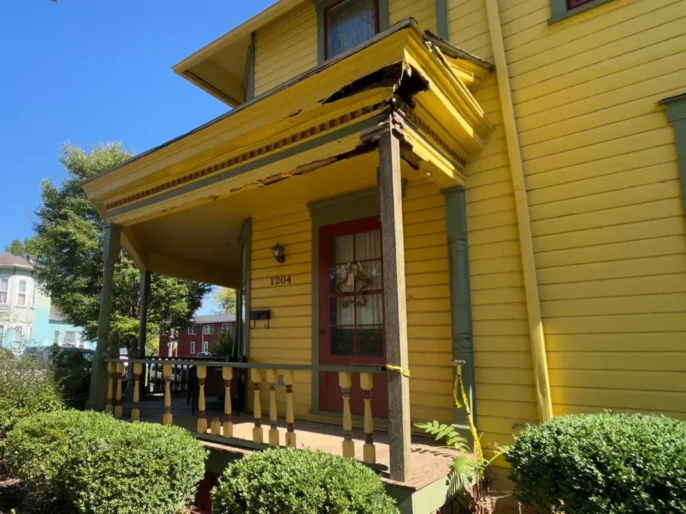 Yellow house with porch damage; red door. Green bushes and blue sky.