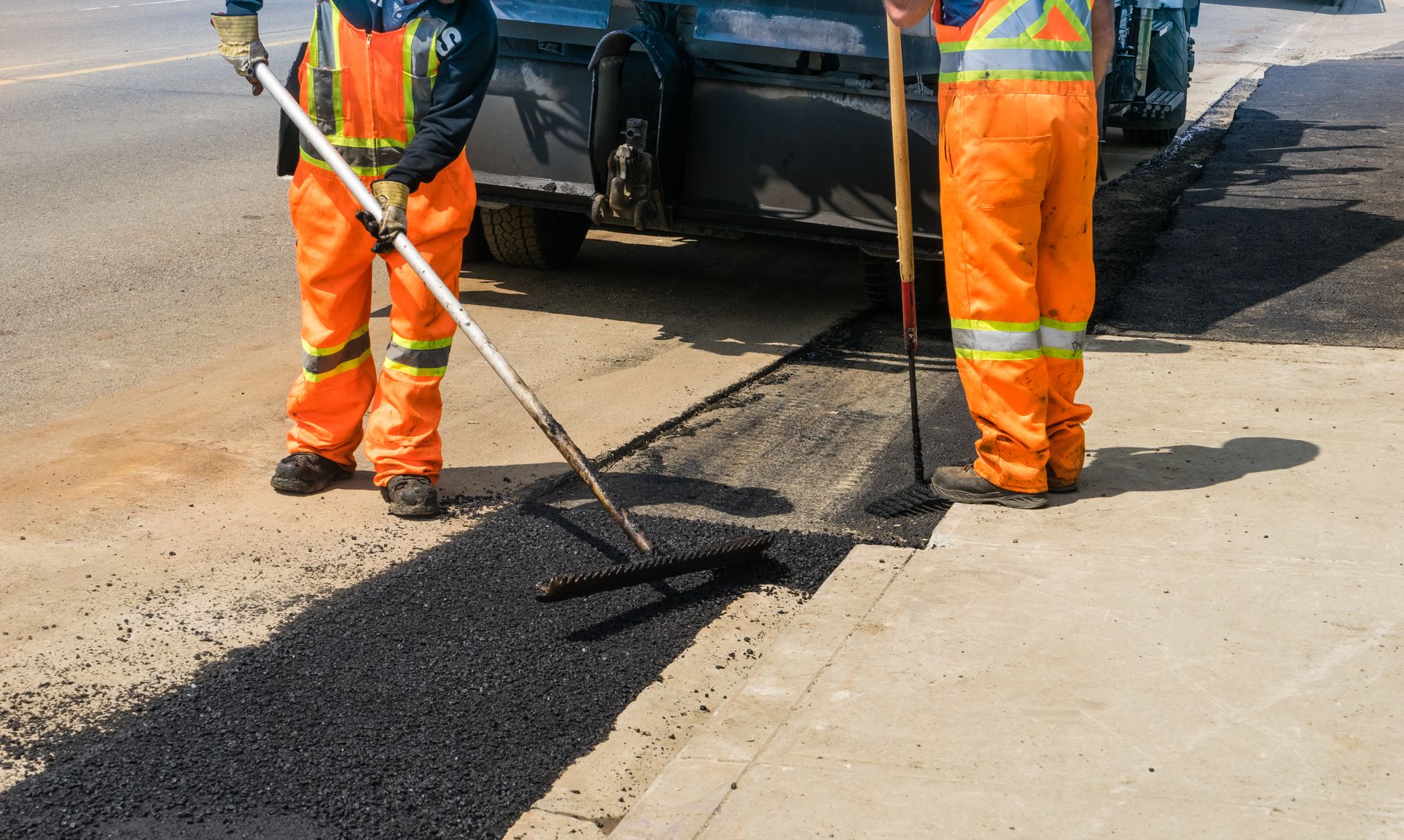 Two workers in bright orange safety gear use tools to spread fresh black asphalt on a road surface.