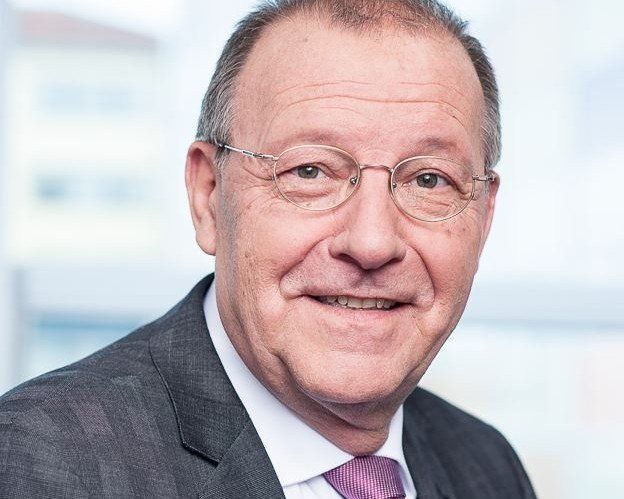 Mann mit Brille und Anzug, lächelnd, in einem Innenraum.