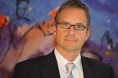 Mann im Anzug und mit Brille, lächelnd, vor einem farbenfrohen abstrakten Gemälde.