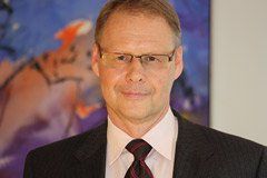 Mann mit Brille und Anzug, lächelnd, steht vor einem farbenfrohen abstrakten Kunstwerk.