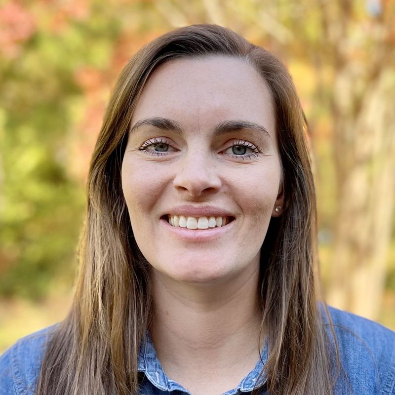 Woman with brown hair smiles at the camera, wearing a blue shirt, with a blurred outdoor background.