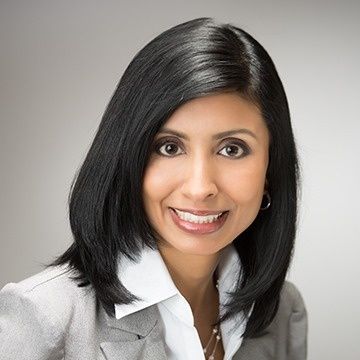 Woman with dark hair smiling, wearing a gray blazer and white shirt, looking at the camera.