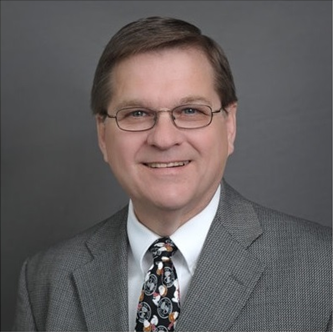 Bald man in a suit smiles at the camera, against a grey backdrop.