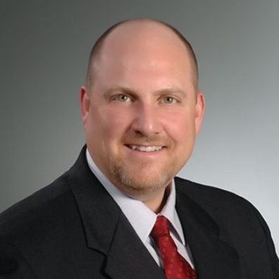 Bald man in a black suit with a red tie, smiling against a gray background.