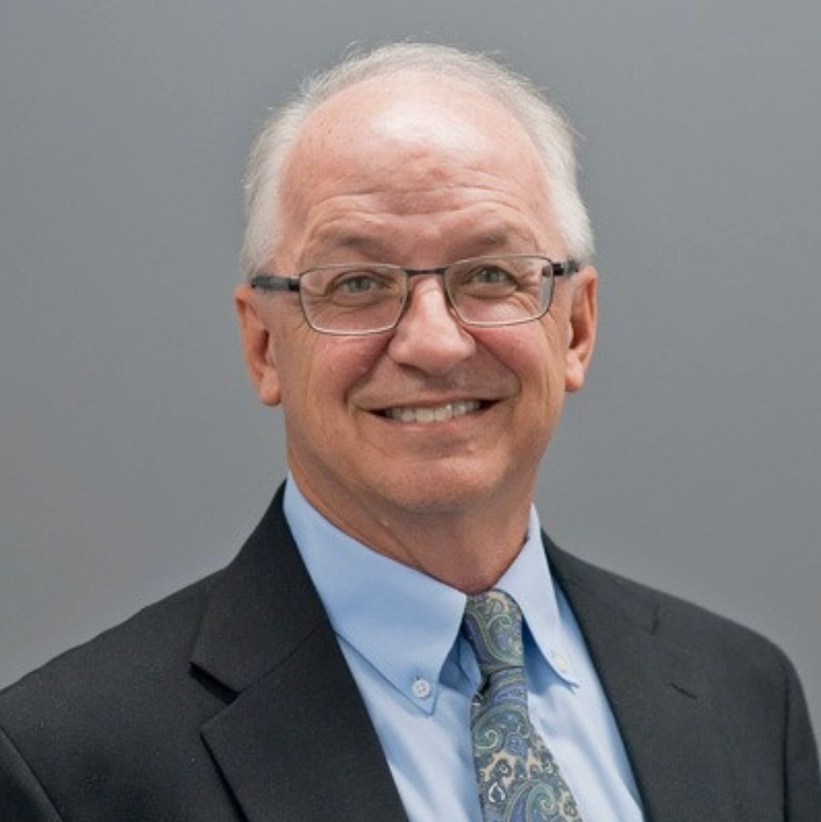 Bald man in a suit smiles at the camera, against a grey backdrop.