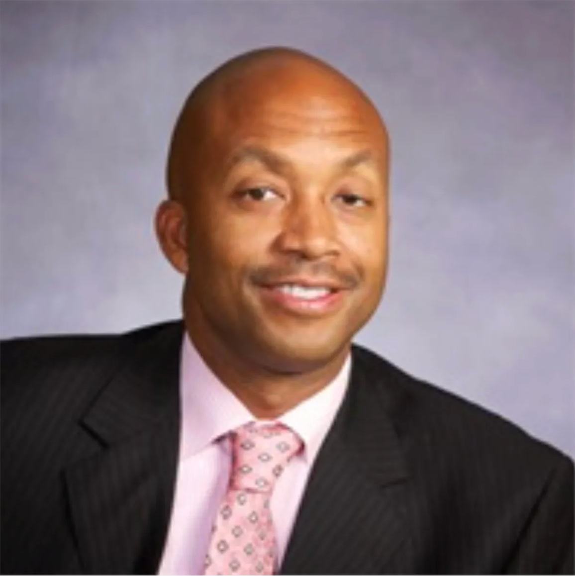 Bald man in a suit smiles at the camera, against a grey backdrop.
