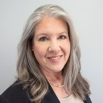 Woman with long, gray hair smiles, wearing a black blazer and pearl necklace, against a white background.