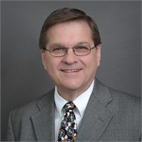 Man in suit and tie smiling, wearing glasses; headshot against gray background.