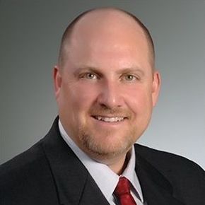 Bald man in a suit smiles at the camera, against a grey backdrop.