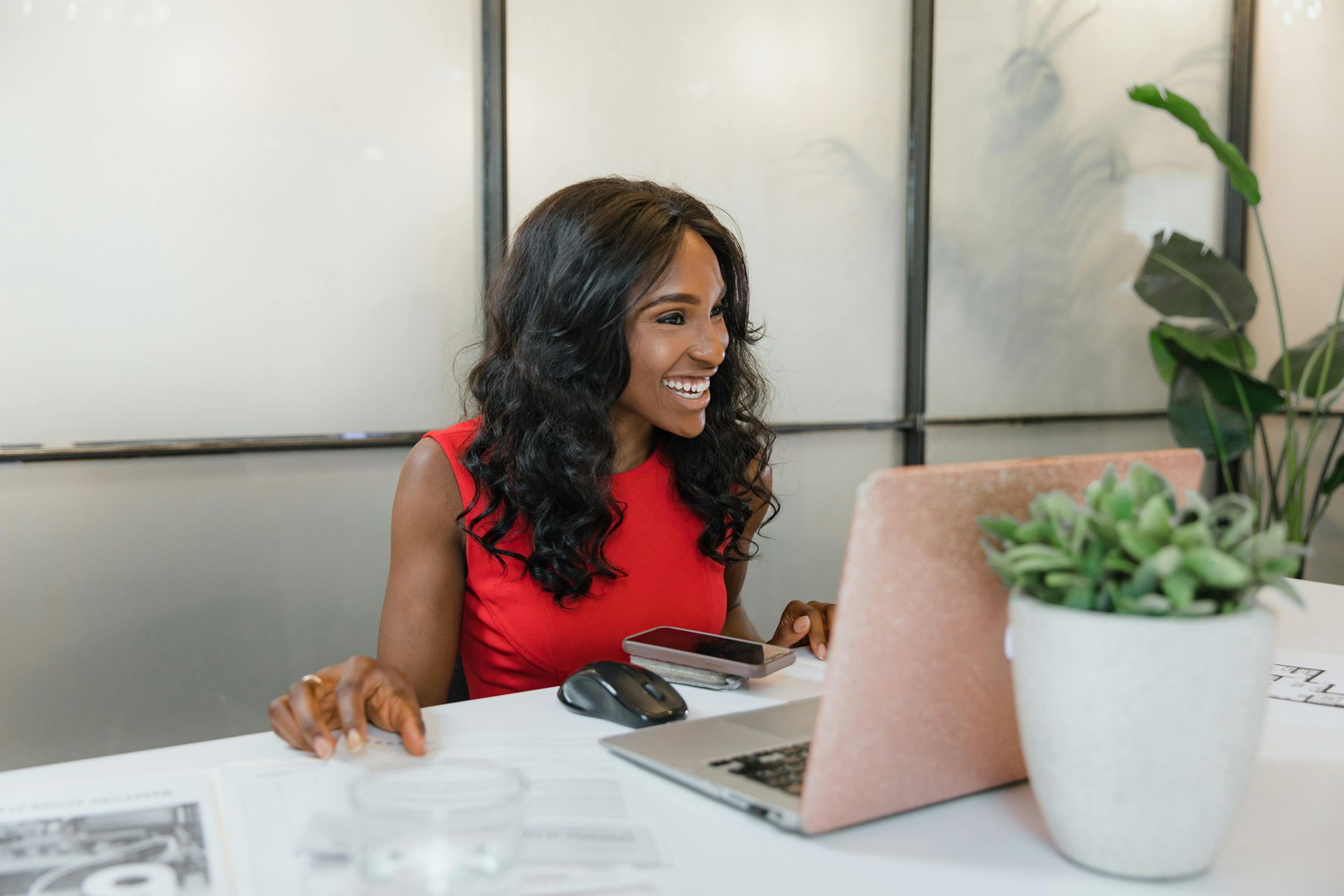 Woman in red dress smiles while working on a laptop at a white desk in an office.