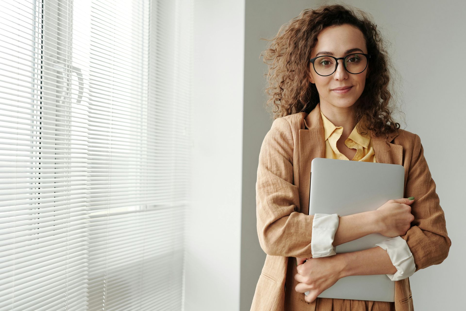 Woman in glasses and a tan blazer, holding a laptop, near a window with blinds.