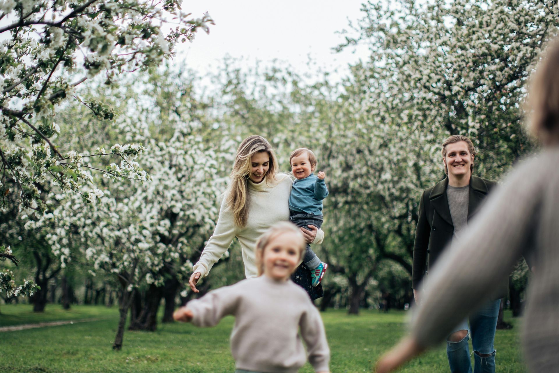 Family in a blooming park; a woman holds a child, two others run toward a man, trees in the background.
