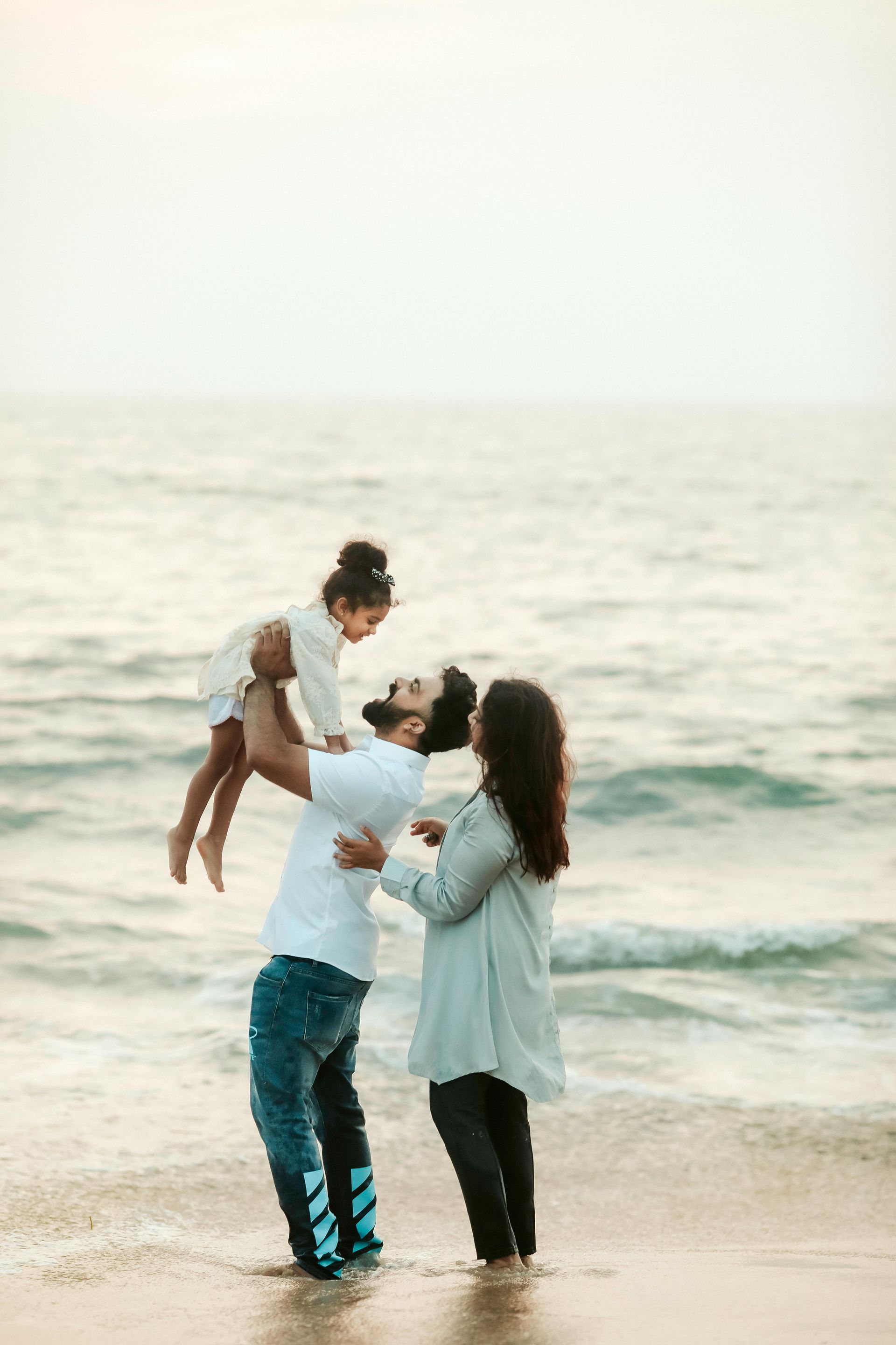 Family at beach: father holding child up, mother watching, gentle waves.