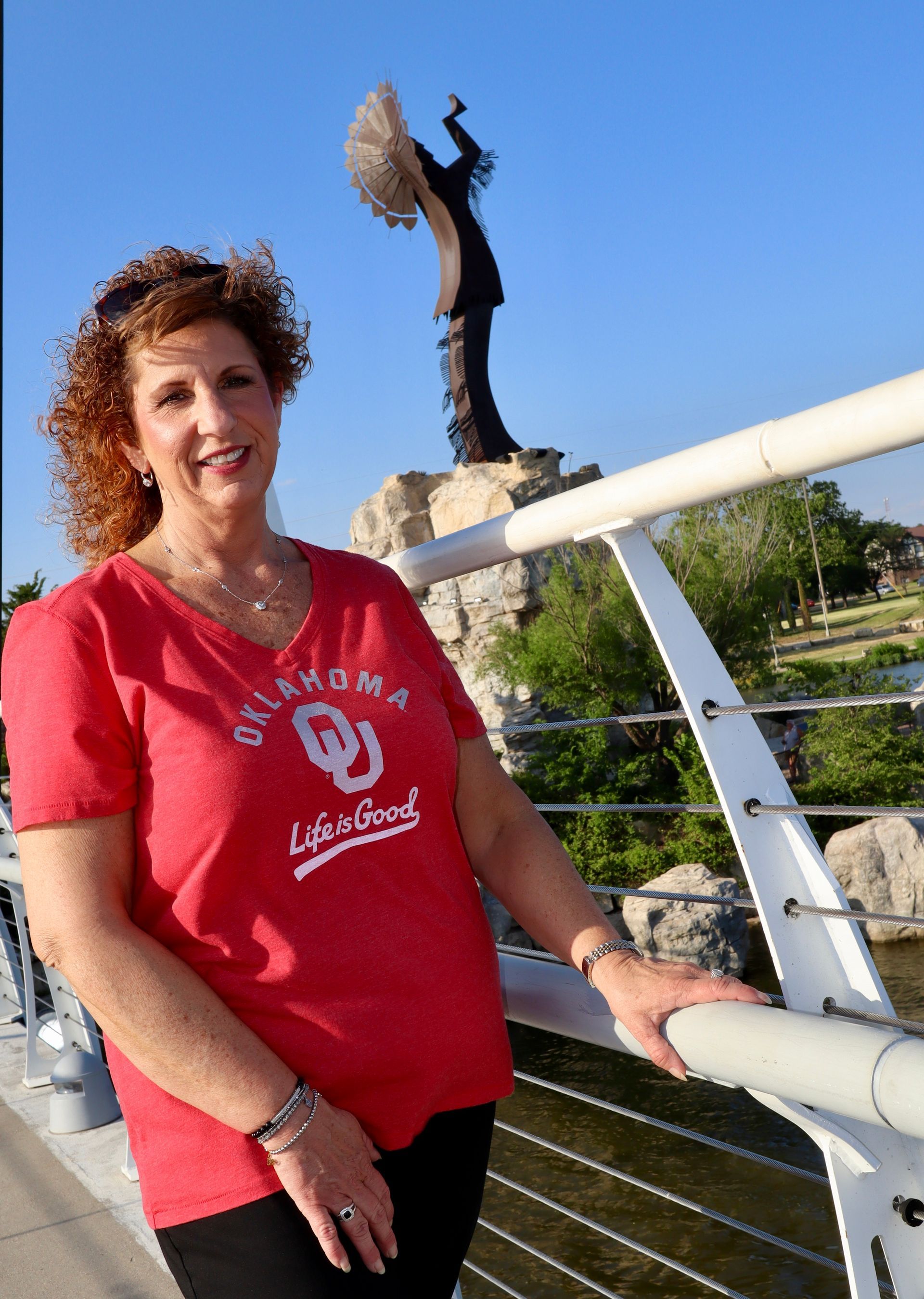 A woman in a red oklahoma shirt is standing on a bridge.