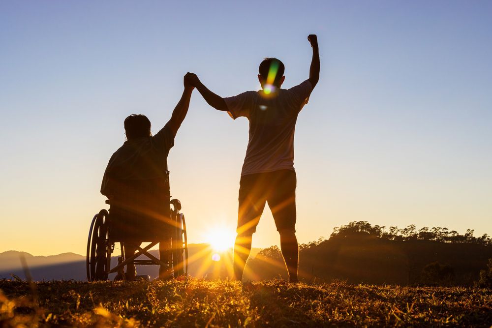 Person in Wheelchair and Standing Person Holding Hands at Sunset in a Field — Coast 2 Coast Disability Support Services in Taree, NSW