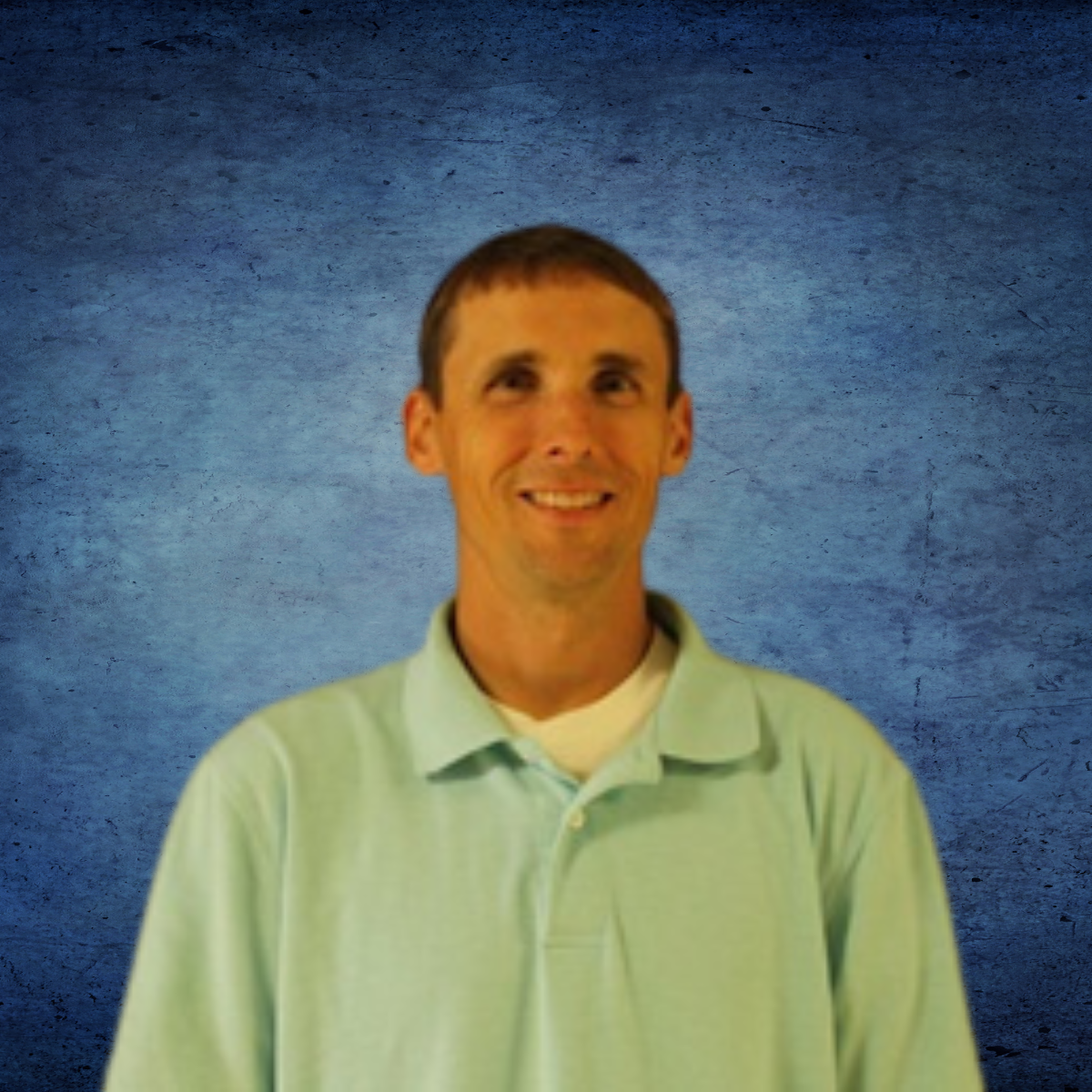 A man in a light blue shirt is smiling in front of a blue background
