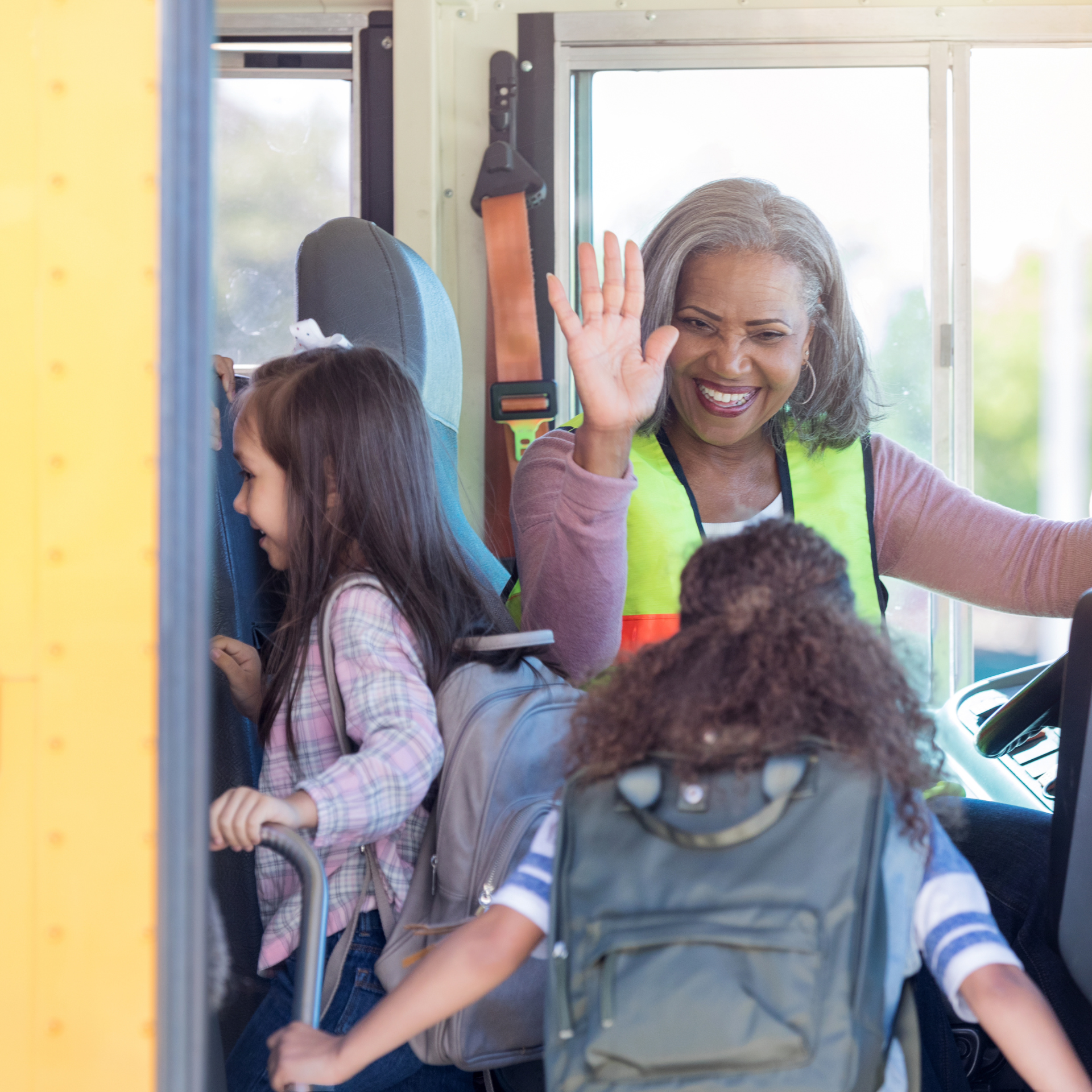 A woman in a yellow vest is waving from the back of a school bus.