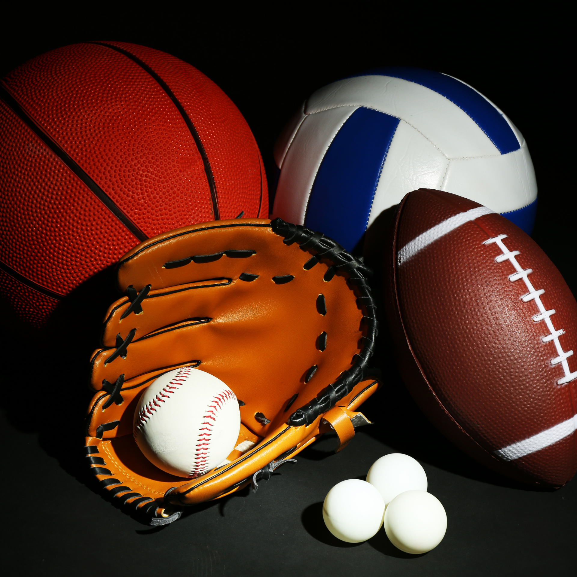 A baseball glove with a baseball in it sits next to a volleyball and a football