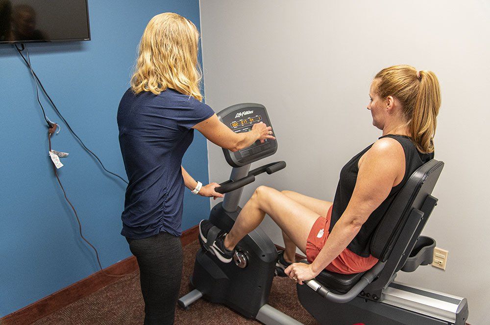 Woman on recumbent bike being assisted by another person in a therapy room.