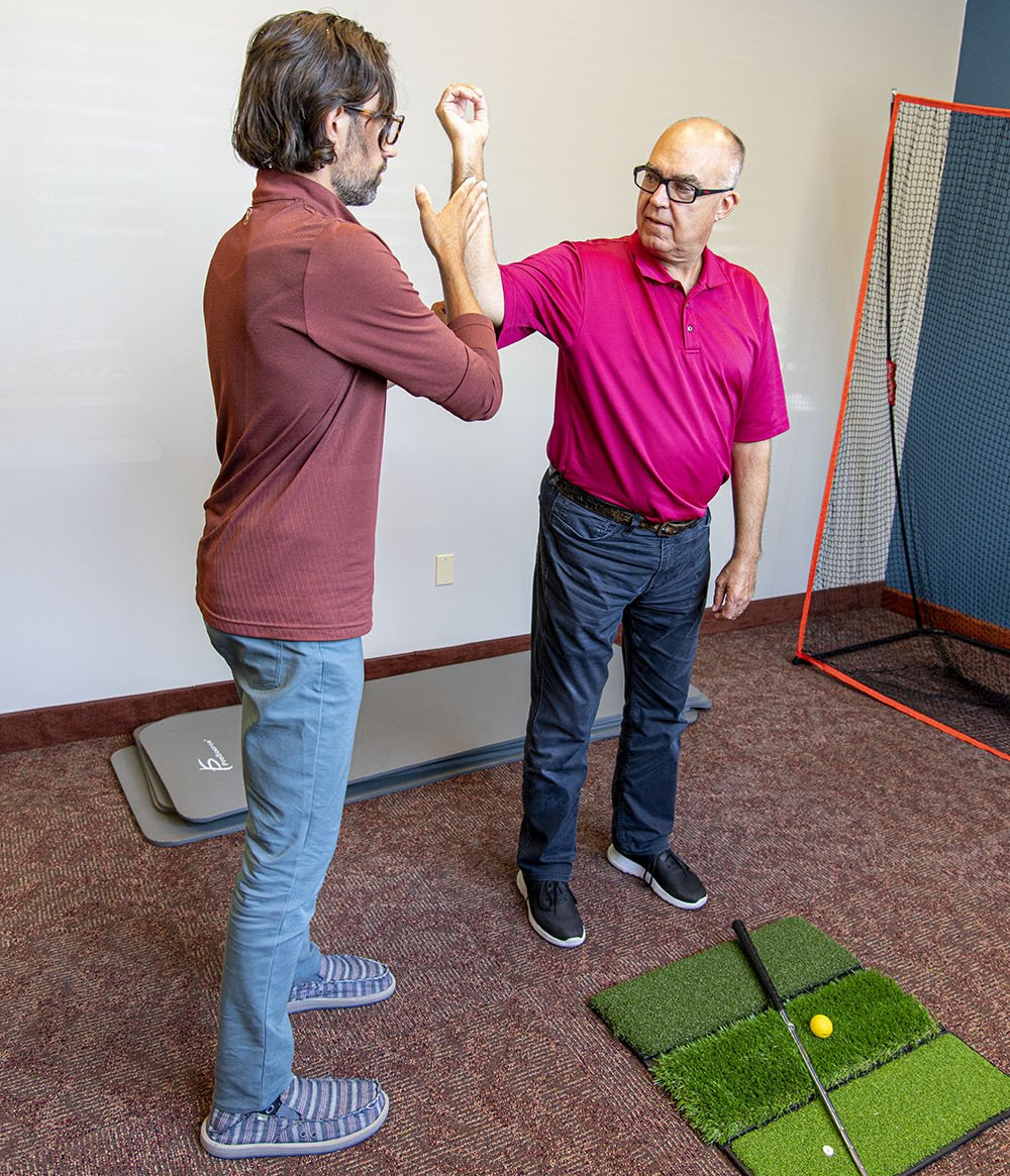 Man in red shirt guiding man's arm during exercise. Golf mat and net in the background. Indoor setting.