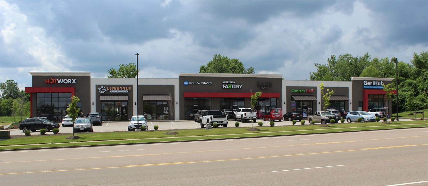 A strip mall with various storefronts, cars parked in front, and a cloudy sky overhead.