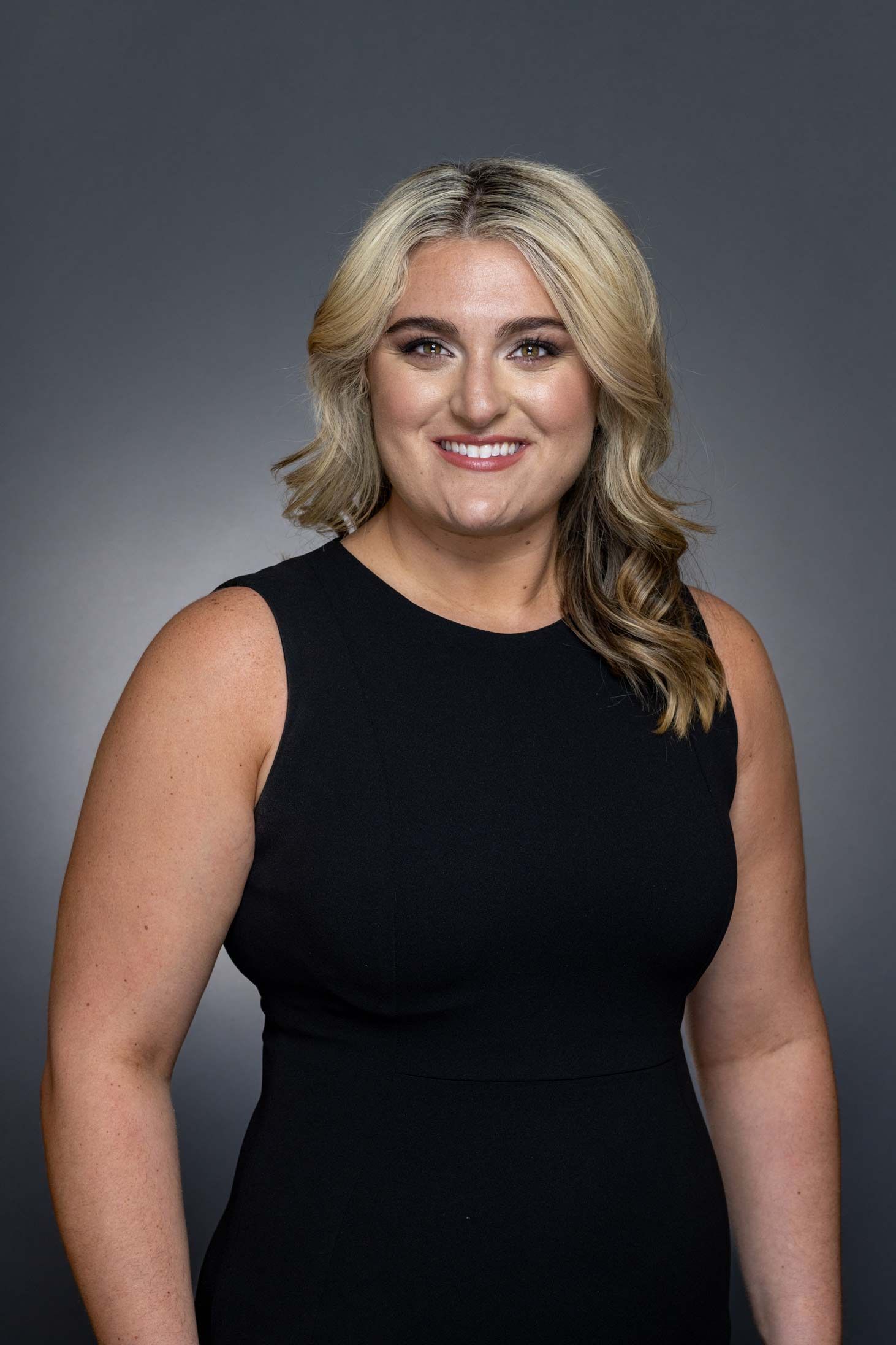 Woman with blonde hair wearing a sleeveless black dress, smiling against a gray backdrop.