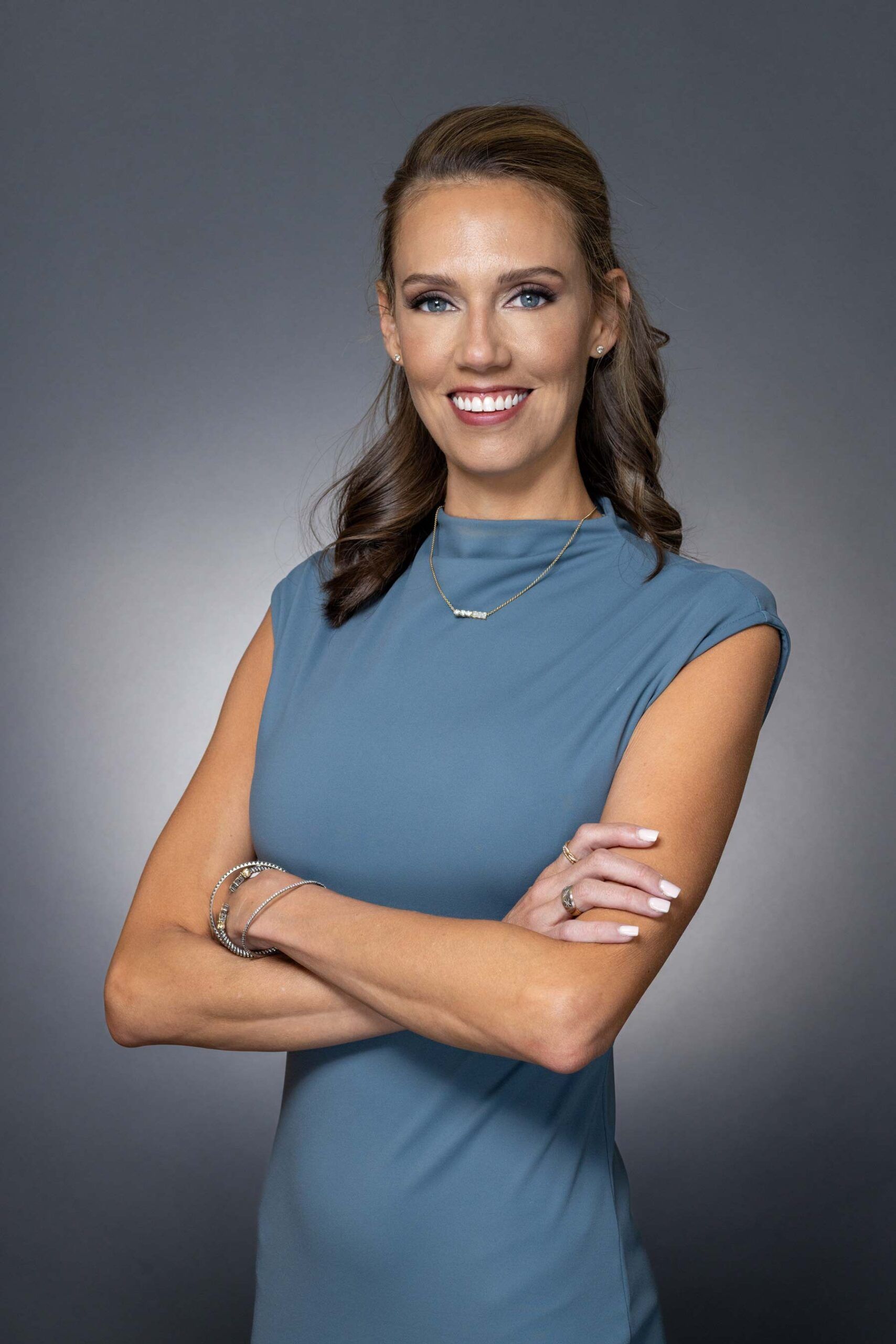 Woman in a teal dress, arms crossed, smiling at the camera against a gray backdrop.