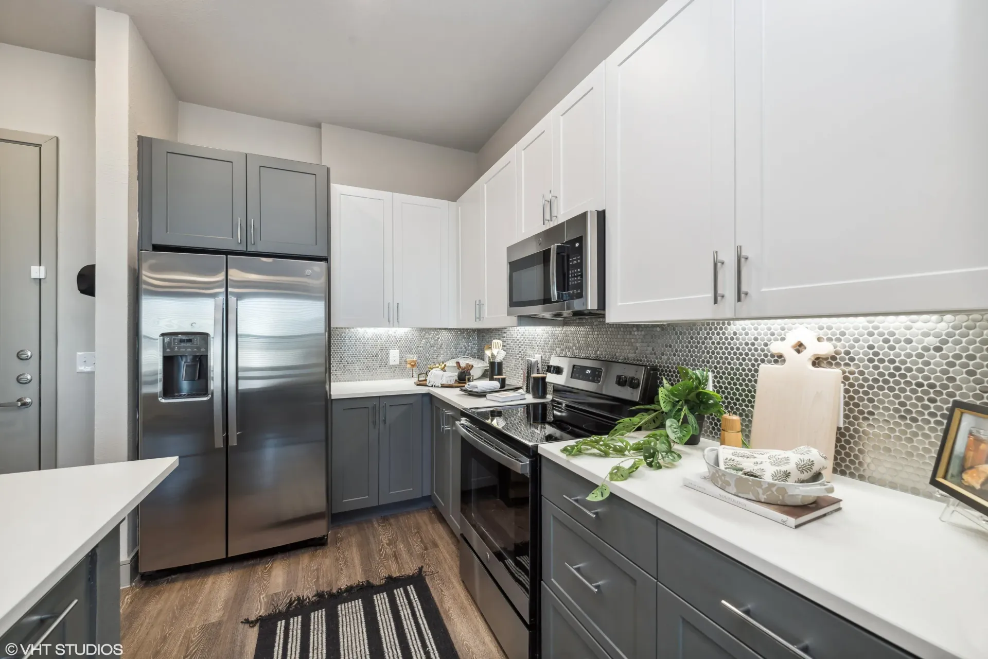 Modern apartment kitchen with stainless steel appliances, white uppers, gray lowers, and a mosaic backsplash.