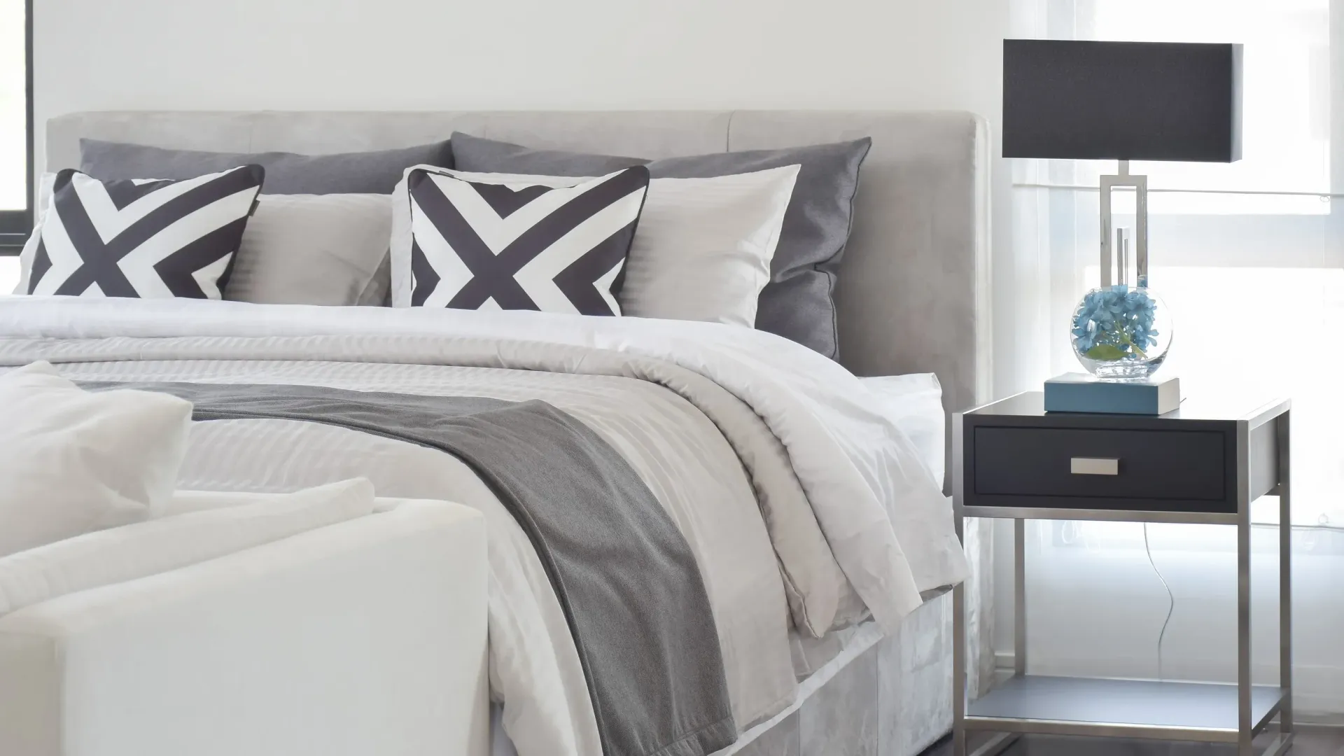 Bedroom interior with a gray upholstered headboard, white bedding, and a nightstand lamp.