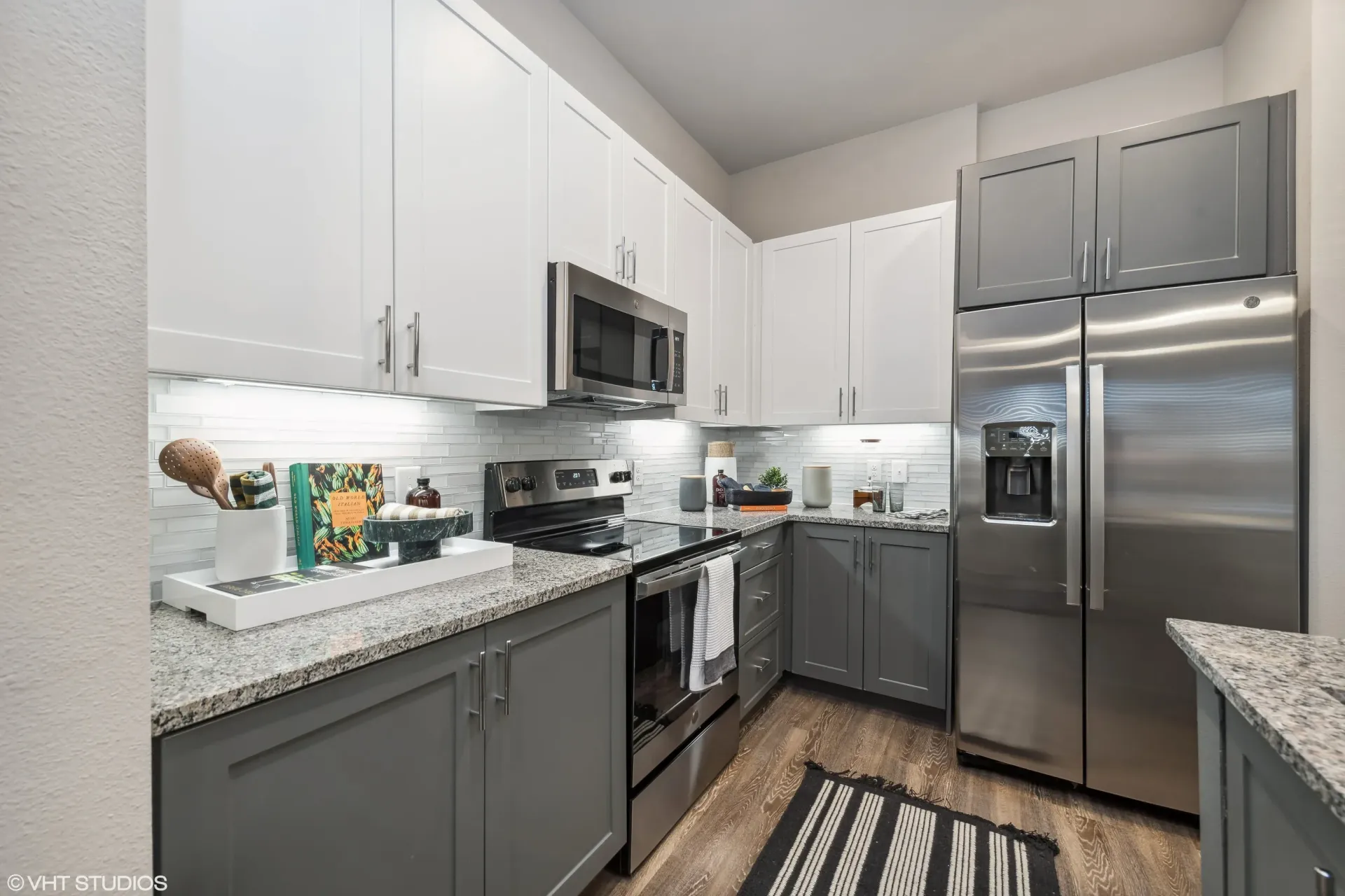 Modern kitchen in apartment with white upper cabinets, gray lower cabinets, granite counters, and stainless appliances.