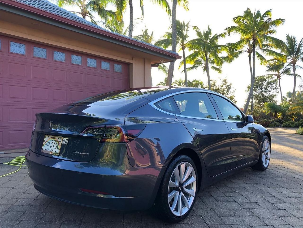 a black tesla model 3 is parked in front of a garage with palm trees in the background