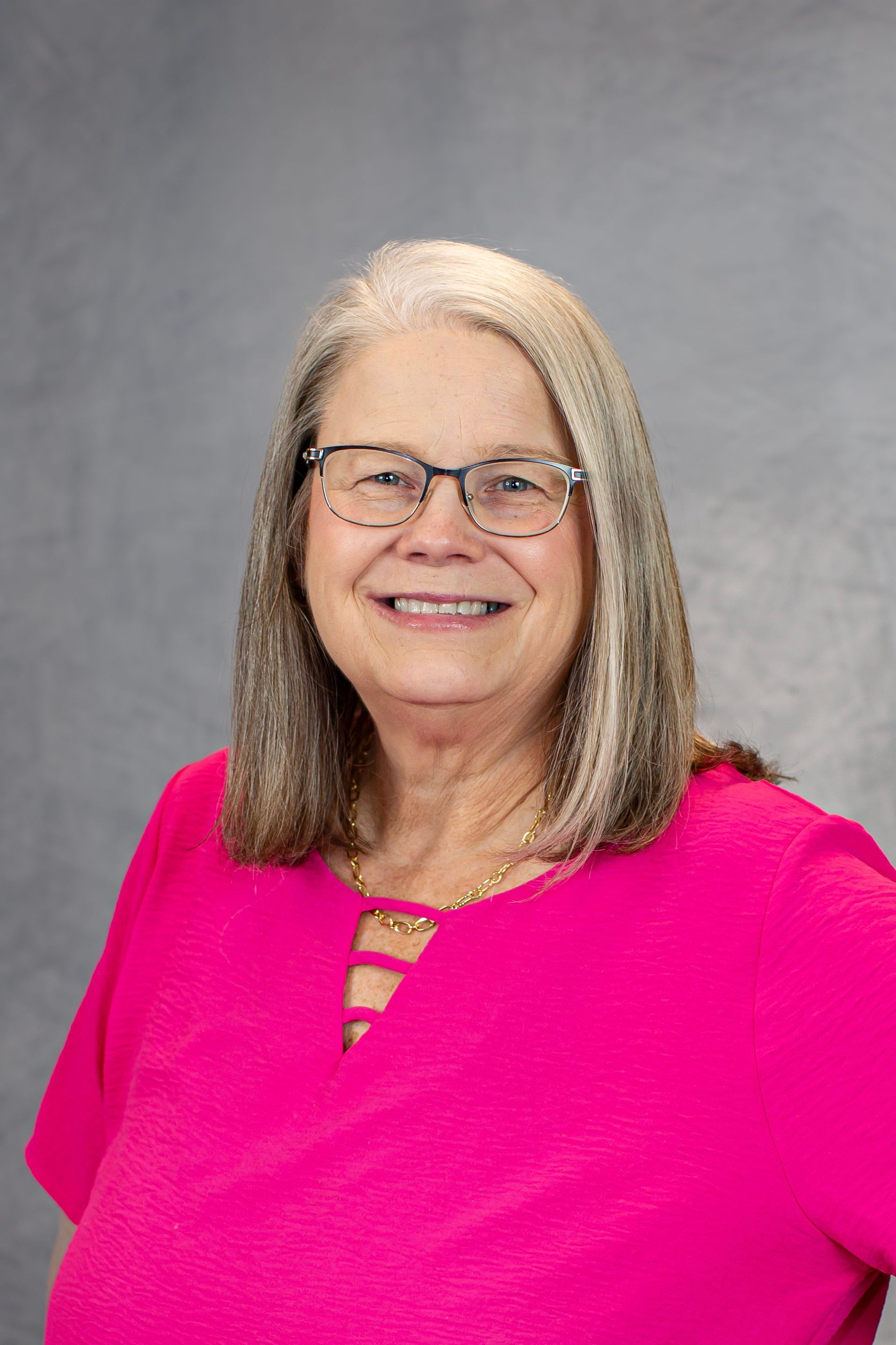 A woman wearing glasses and a pink shirt is smiling for the camera.