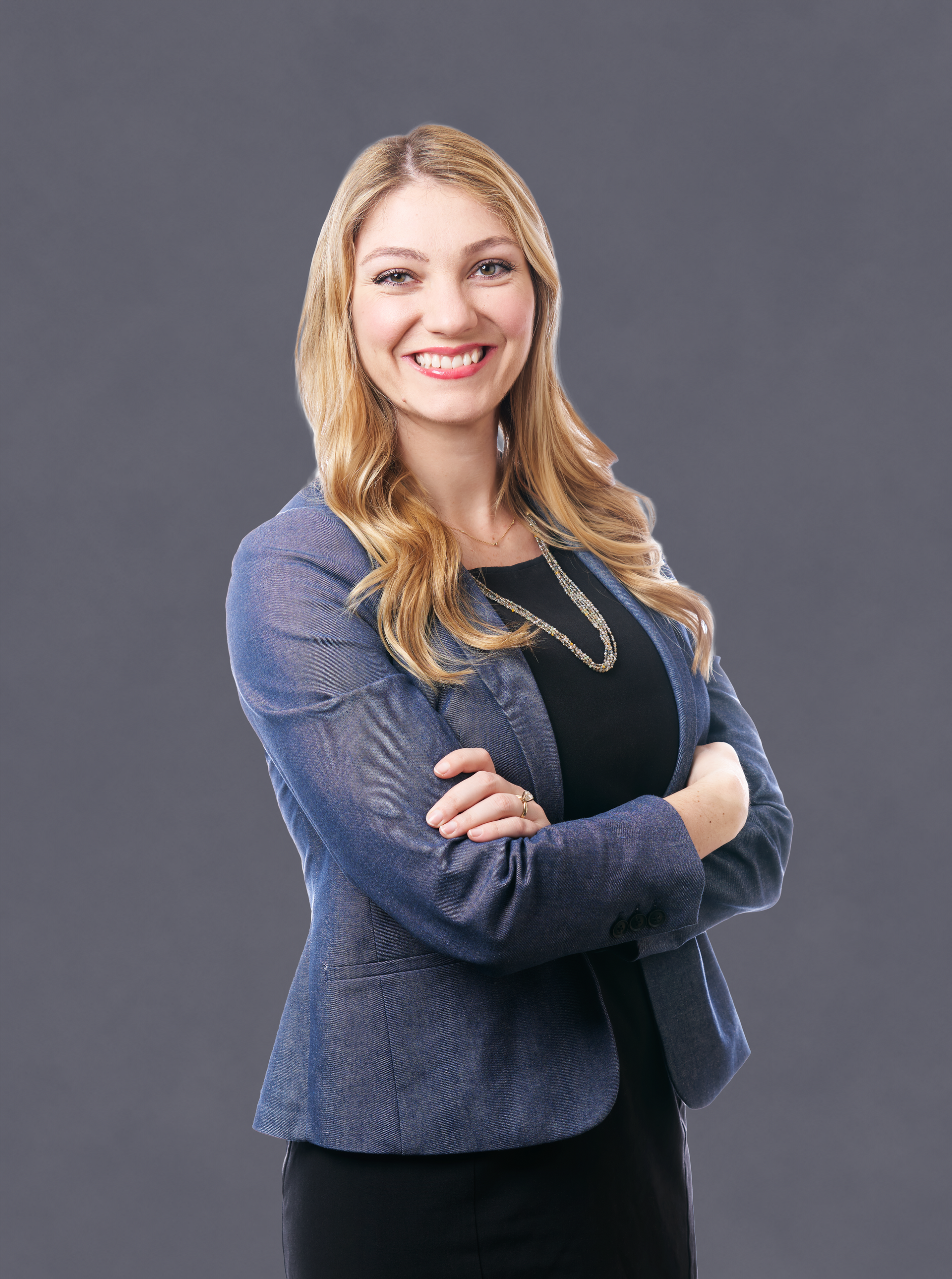 A woman in a blue jacket and black dress is standing with her arms crossed and smiling.