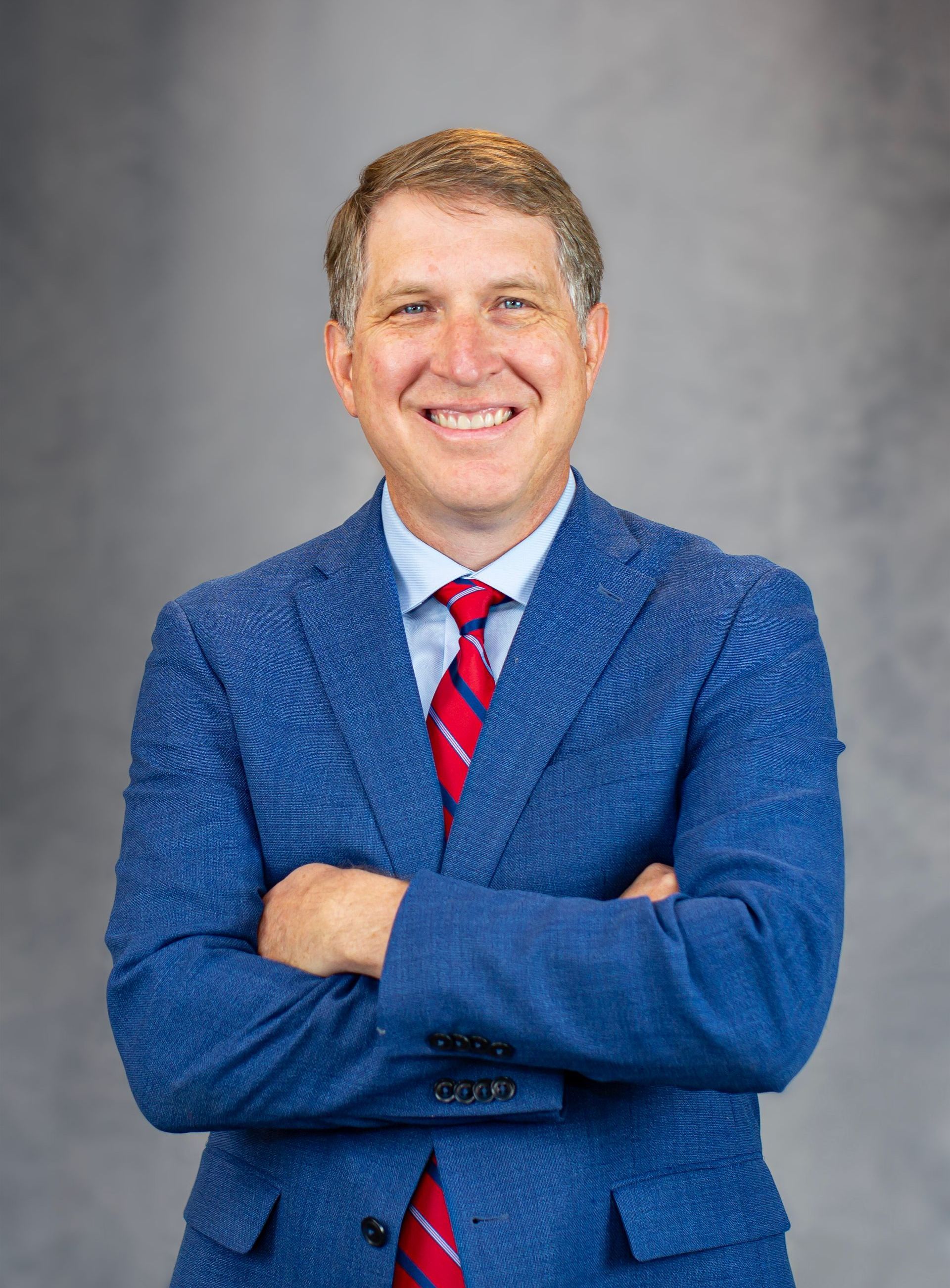 A man in a blue suit and red tie is smiling with his arms crossed.