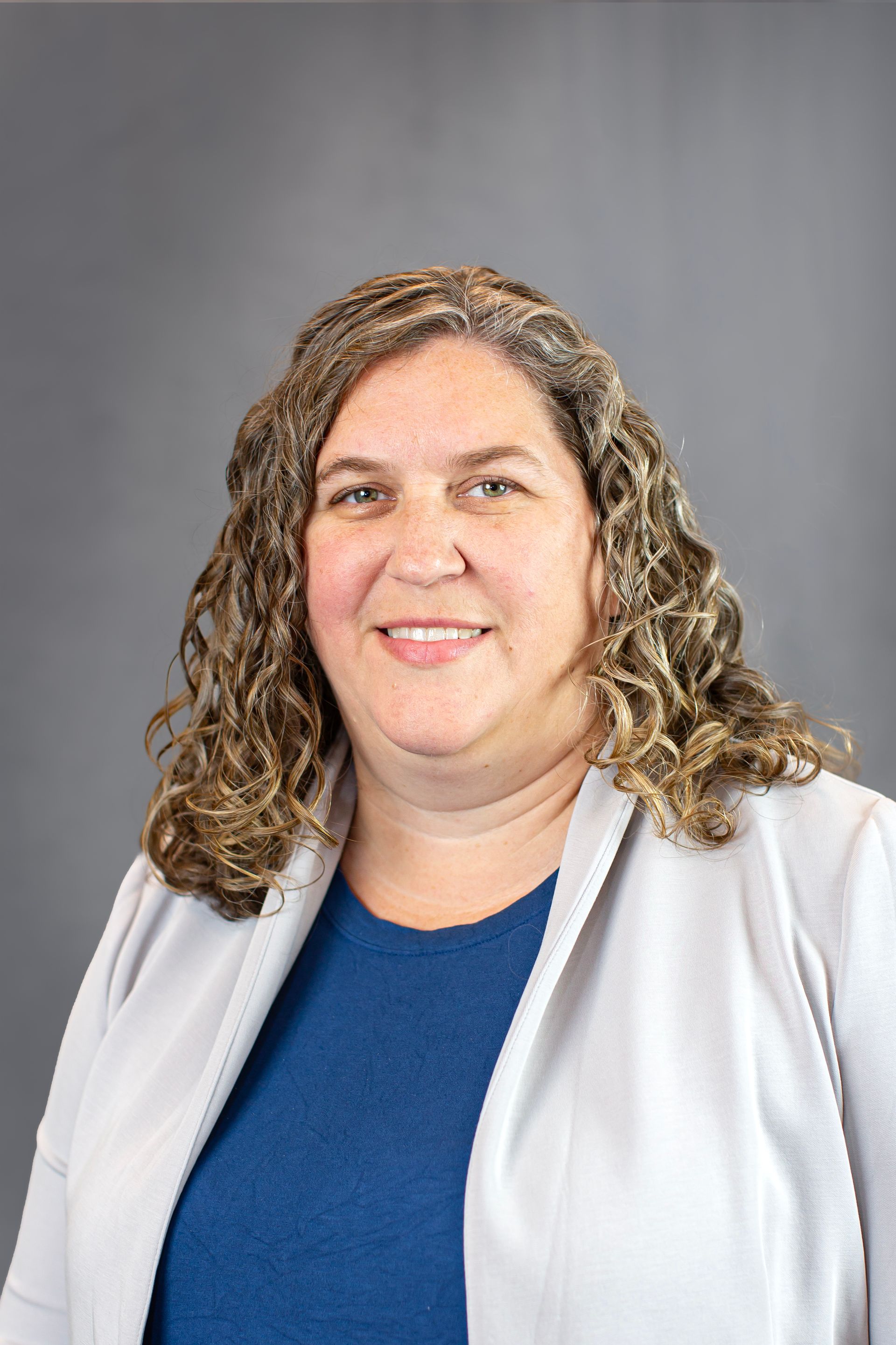A woman with curly hair is wearing a blue shirt and a gray jacket.