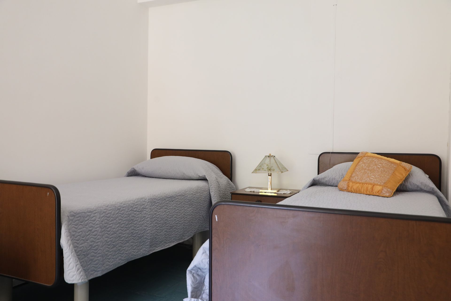 Due letti singoli in una stanza, con un comodino e una lampada in mezzo. Uno dei letti ha un cuscino arancione.