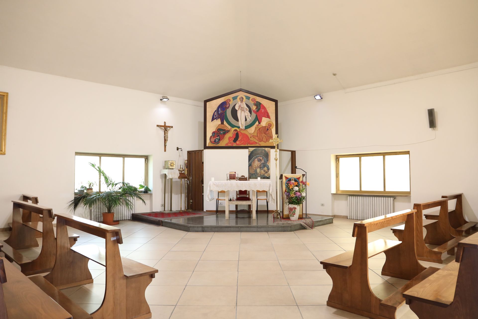 Interno di una cappella con panche in legno, altare, croce e colorate opere d'arte religiose.