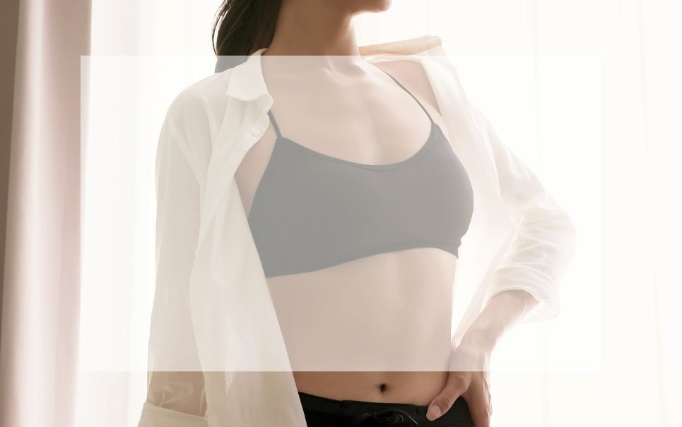 A woman wearing a bra and a white shirt is standing in front of a window.