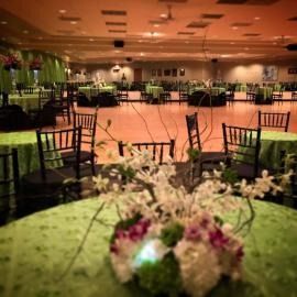 Empty banquet hall with green tablecloths, black chairs, and floral centerpieces under warm lighting