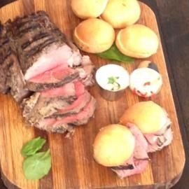 Sliced roast beef with bread rolls and sliced mozzarella on a wooden cutting board, garnished with basil.