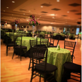 Banquet hall with round tables, black chairs, and green tablecloths under warm lighting
