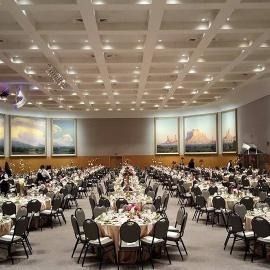 Banquet hall with round tables and chairs set for a large formal event, under a coffered ceiling and landscape art.