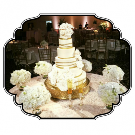 Elegant white tiered wedding cake on a gold stand, surrounded by white floral arrangements at a reception.
