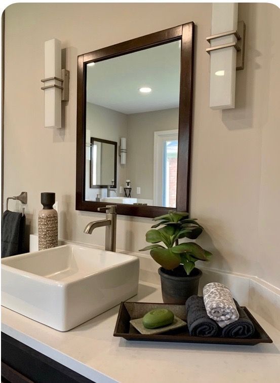 A modern bathroom vanity with a square white sink, dark wood mirror, wall sconces, and a small plant on a tray.