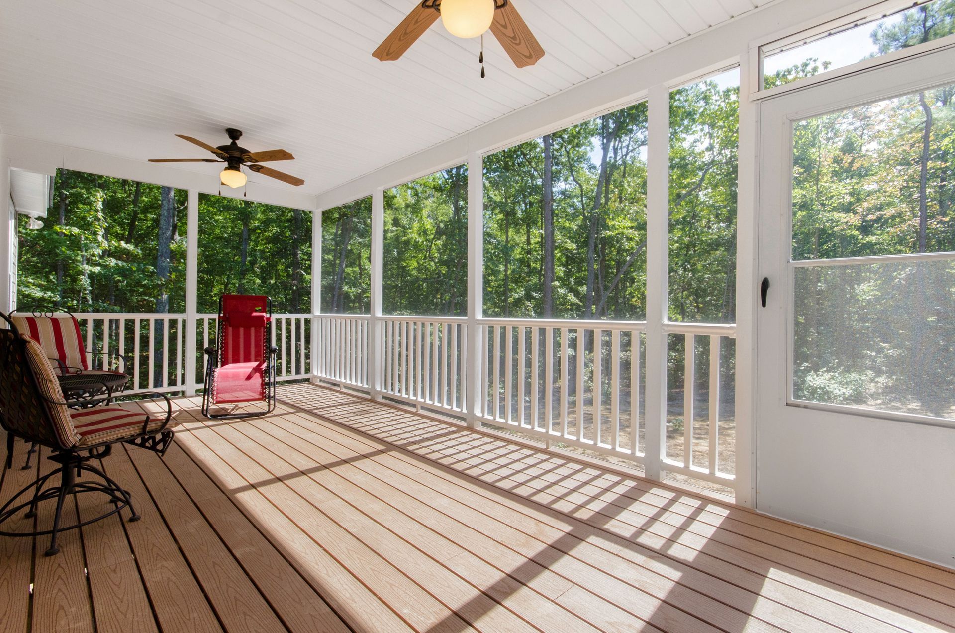 Sunlit screened porch with ceiling fans, red chair, wicker chairs, and white railing overlooking trees